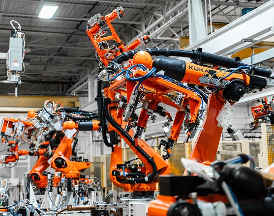 Several orange KUKA industrial robots equipped with grippers are operating along an automated production line in an industrial hall, handling workpieces as part of the foundry automation process. In the background, additional robots, machinery, and the hall’s structure can be seen.