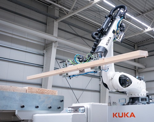 An industrial robot arm in a factory hall, gripping and positioning a long wooden beam horizontally, mounted on an automated production line. The word “KUKA” is clearly visible on the white robot arm; in the background are steel beams, lighting, and other machinery typical of an industrial setting.