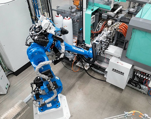 A blue industrial robot arm bearing the inscription “Yaskawa” is operating on an “Arburg” production machine in an automated manufacturing environment.