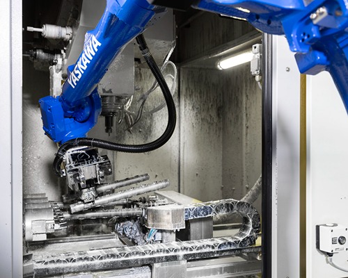 A blue industrial robot arm bearing the inscription “Yaskawa” is machining a metal component inside a closed machine tool. Metal shavings and coolant are visible in the work area.