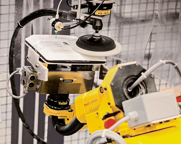 A yellow FANUC industrial robot is equipped with a rotating polishing tool and automatically processes the surface of a clamped workpiece within an industrial facility. Hoses, sensors, and safety guards for the polishing process are visible.