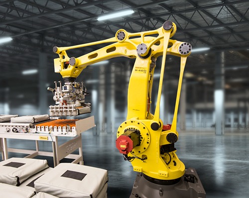A yellow FANUC industrial robot equipped with a gripper system is located in an industrial hall, lifting several bags from a conveyor or loading station. Next to the robot, stacked bags, a feeding unit, and the hall structure are visible in the background.